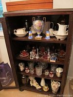 Full view of wooden cabinet with multiple shelves displaying a variety of glassware, vases, bells, pewter cups, and figurines including shoe figurine and ceramic vases.