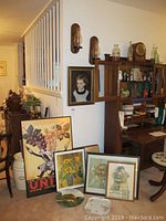 Overall view showing framed wall art leaning against wall and bookcase, decorative plates on floor, and wooden wall sconces mounted on wall.