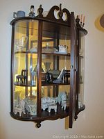 Full view of the wooden wall cabinet with glass curved doors showing interior contents of miniature furniture and glassware.