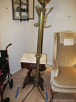 Full view of brass-colored metal coat rack and wooden side table with marble top, next to beige upholstered chair and mobility walker.