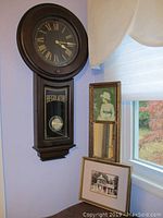 Regulator wall clock mounted on wall with visible Roman numeral dial and pendulum behind glass labeled 'Regulator'. Below the clock are three pieces of framed wall art.