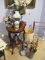 Photo showing assortment of decorative items on small wooden table and floor, including two floral lamps, faux flower arrangements in vases, wicker basket, lantern, alarm clock, and cylindrical lampshade.