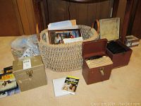 Photo showing a woven basket on the floor with two cardboard record boxes and a third beige storage box, along with a plastic bag of additional media items and DVDs arranged nearby.