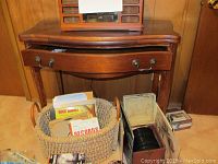 Front view of brown wooden table with a slightly curved drawer open. A basket containing vinyl records is on the floor in front; the table rests against a wood paneled wall.