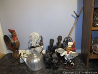 Photo showing entire lot with three wooden roosters (large brown, medium white, small white swan), two African figurines, chef figurine holding trays, and large metal kettle with black handle.