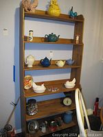 Full view of wooden shelf containing various ceramic teapots in blue, green, white, and yellow, a ceramic pitcher, salt and pepper shakers, glass jar with cookie cutters, Columbia scale, and assorted vintage kitchen items.