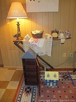 Photo showing lamp, small woven basket on ironing board, bird figurine, doll, miniature flag, hand towels and doilies, decorative wood houses on rug and shelf