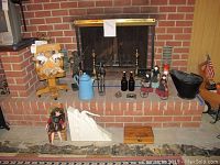 Photo showing arrangement of wood doll chair, plush bear, metal figurines, kettle, brown bottles, plush dolls on wagon, ash can, wooden box, and white carving in front of fireplace.