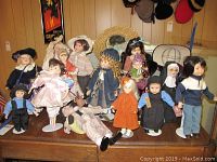 Collection of 13 porcelain dolls displayed on wooden table against wood-paneled wall with hats hanging above.