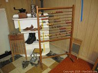 Wide shot displaying most lot items: vintage shoes, shoe stretchers, wooden abacus, two washboards, glass canister with buttons, metal cobbler tool, two metal grinders, empty cigar box, all arranged against wood paneled wall and checkered floor.
