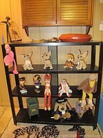 Three-tier black shelf with assorted figurines and plush animals including bobbleheads, carved cats, ceramic dogs, plush flamingo and turkey