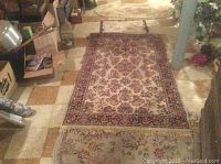 View of the large burgundy and cream floral pattern area rug with smaller throw rugs lined behind it in a room with tiled floor.
