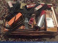 Photo showing scattered Tyco train track pieces, some curved, some straight, with a red and white Santa Fe locomotive and other freight cars on top of a cardboard box labeled 'Electric Train'. Various small parts for assembly visible.