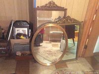 Three vintage wood framed mirrors placed leaning against a wall. Oval mirror with gold frame front and center, rectangular mirrors behind.