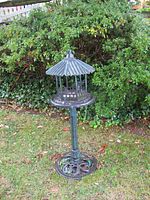 Metal bird feeder on stand with pagoda-style roof seen from an angle, standing on grass near shrubbery.