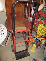 Front view of metal dolly with attached white bungee cord, red base plate, black wheels, placed in front of rusty metal wheelbarrow with wood handles and reddish basin.