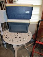 Two Igloo coolers stacked on a round plastic patio table with floral cutout design. Background shows part of metal folding tables.