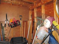 Photo showing various yard tools hanging or standing inside a wooden shed including rakes, a snow shovel, and a scarecrow decoration.