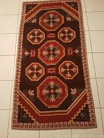 Full view of the rectangular hand knotted wool rug showing geometric and floral medallion patterns with a red border on a dark brown background.
