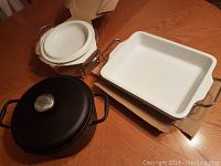 View of three cookware items: cast iron pot with lid, white ceramic round pot with lid and metal stand, white ceramic square pan with metal stand on a wooden table.