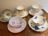 Five vintage bone china teacups and saucers displayed on a wooden surface. Assorted floral patterns and gold trims visible.