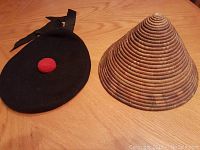 Photo of both hats side by side on wooden surface: black military beret with red pom, conical African woven straw hat.
