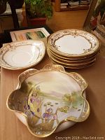 Photo showing the scalloped Nippon porcelain bowl with hand-painted irises and a scenic background, alongside a stack of 8 matching plates with a gold and brown patterned rim.