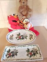 Full view of two oval Christmas themed serving trays with holly and berry decoration, surrounded by a beige teddy bear, red Elmo and Tigger plush toys on tiled background.