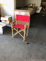 Red directors chair with bamboo-style wooden frame, armrests, and red fabric seat and backrest, photographed indoors next to cardboard boxes.