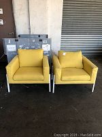 Frontal view of pair of yellow fabric armchairs with white metal legs on a concrete floor in warehouse setting.