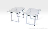 Pair of side tables with clear glass tops and chrome rectangular leg frames photographed from an angled top-front view.