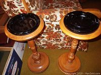 Two wooden ashtray stands side by side with circular black ceramic ashtrays on top, next to a floral upholstered couch.