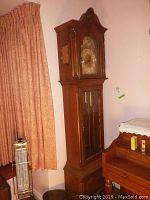 Full height view of wooden grandfather clock with carved pediment and glass front door, positioned near curtains and heater