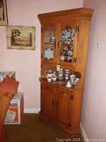 Maple corner china cabinet with two glass doors on top showcasing red, white, and black patterned china, and two wood panel doors below with brass handles. Cabinet has a decorative scalloped top trim and is positioned in a corner with pink walls.