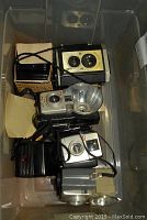 Box of six vintage cameras including Baby Brownie models and others with attached straps and mounts inside a plastic container