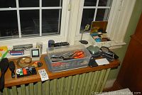 Wide view of assorted items including bin with scissors, magnifying glasses, brushes, brass tray, magnifying stand, and more on windowsill and radiator cover.