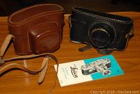 Two leather camera cases (black and brown) alongside Leica instruction booklet on wood surface