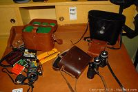 Three sets of binoculars in leather cases laid on a wooden surface along with a brown leather camera bag and various camera accessories including exposure meters and filters.
