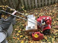 Red Mantis garden tiller with chrome handle assembly, sealed Aerator/Dethatcher combo box and two red plastic jerry cans positioned on leaf-covered grass