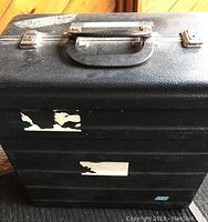 Closed black hard carrying case showing worn labels and handle.