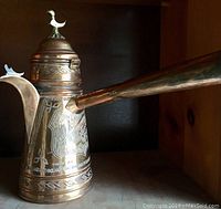 Side view of vintage copper pitcher with long spout and bird finial on the lid, detailed etched designs visible.