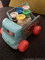 Front angle view of vintage Fisher Price milk wagon showing light blue cab, red wheels, and wooden side panels with illustrations.