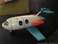 Front and side view of vintage white plastic toy airplane with blue wings, orange engines, black wheels, and blue windshield on a couch.
