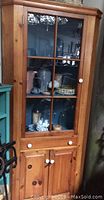 Front and angled view of a solid pine corner cabinet with glass upper door showing two interior shelves with various decorative items.