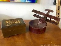 Photo showing both music boxes: a wooden rectangular box with floral inlay and a metal biplane music box.