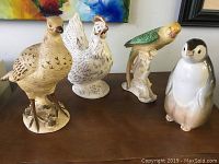 Four vintage ceramic bird figurines including a pheasant, rooster, parrot, and penguin chick, arranged on wooden surface with colorful abstract art background.