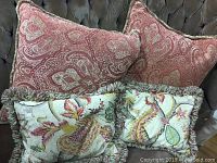 Four down filled cushions on a dark upholstered surface; two large red paisley 22" square cushions and two smaller rectangular floral cushions with moss fringe.