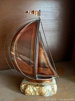 Full view of the copper sail boat sculpture mounted on stone base, showing metal framing and twisted wire rigging.