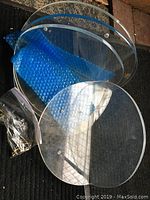 Three round clear acrylic/Lucite shelves stacked or overlapping, shown with blue protective foam and bag of mounting hardware on black carpet.