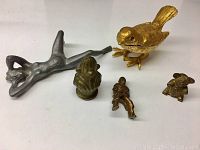 Five miniature metal figures including a reclining nude woman, a bird, frog, soldier, and dog/bear figure on a white surface.